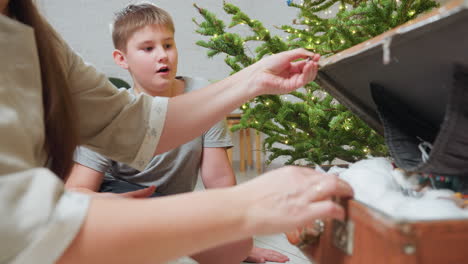Partial view of woman opening box in cozy indoor setting beside Christmas tree with glowing lights, young boy sitting nearby with surprised expression, warm festive holiday moment