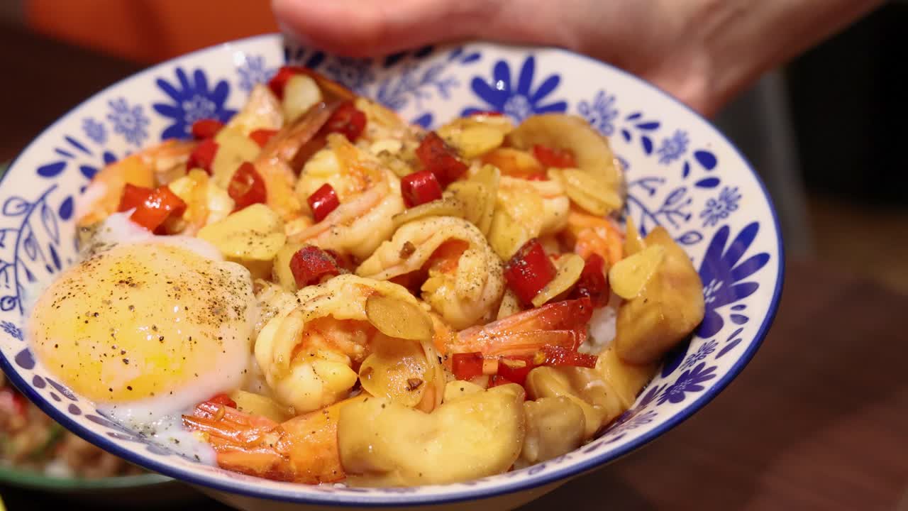 A hand holds a vibrant Thai rice meal with shrimp, runny egg, garlic, chili, and vegetables in a decorative bowl under warm indoor lighting