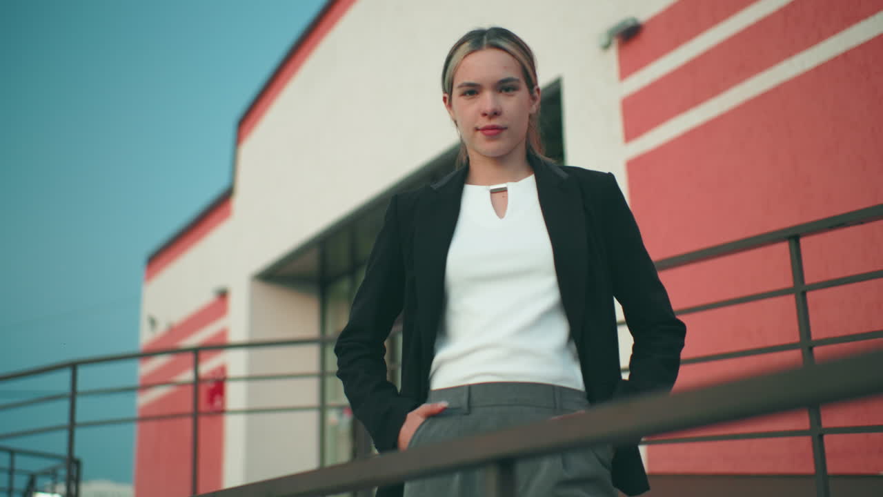 Confident lady posing with warm smile beside iron railing in front of office building featuring striped walls under clear sky, hands in pockets, exuding elegance and professionalism in urban setting