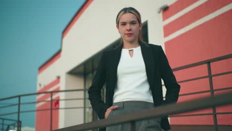 confident lady posing with warm smile beside iron railing in front of office building featuring striped walls under clear sky, hands in pockets, exuding elegance and professionalism in urban setting