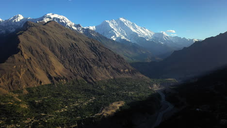 Epic-drone-shot-of-the-sun-shining-in-the-valley,-Passu-Cones-in-Hunza-Pakistan,-snow-covered-mountain-peaks-with-steep-cliffs,-high-wide-aerial-shot