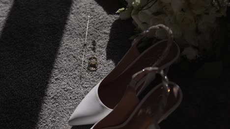 white high-heeled shoes, wedding rings, and necklace on a carpeted floor in sunlight