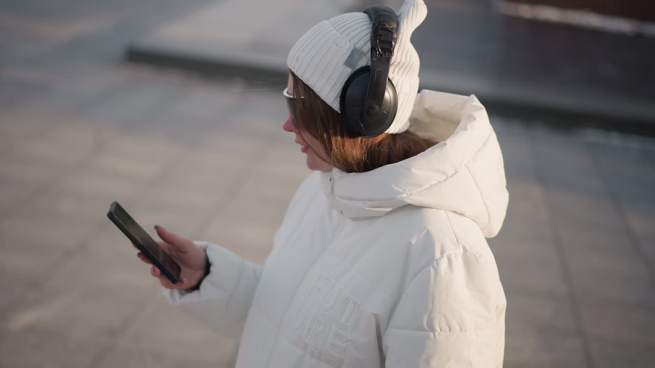 Side view girl sliding through her phone while turning her body wearing tinted goggles headphones and winter coat on snowy urban plaza under soft golden hour light with subtle motion