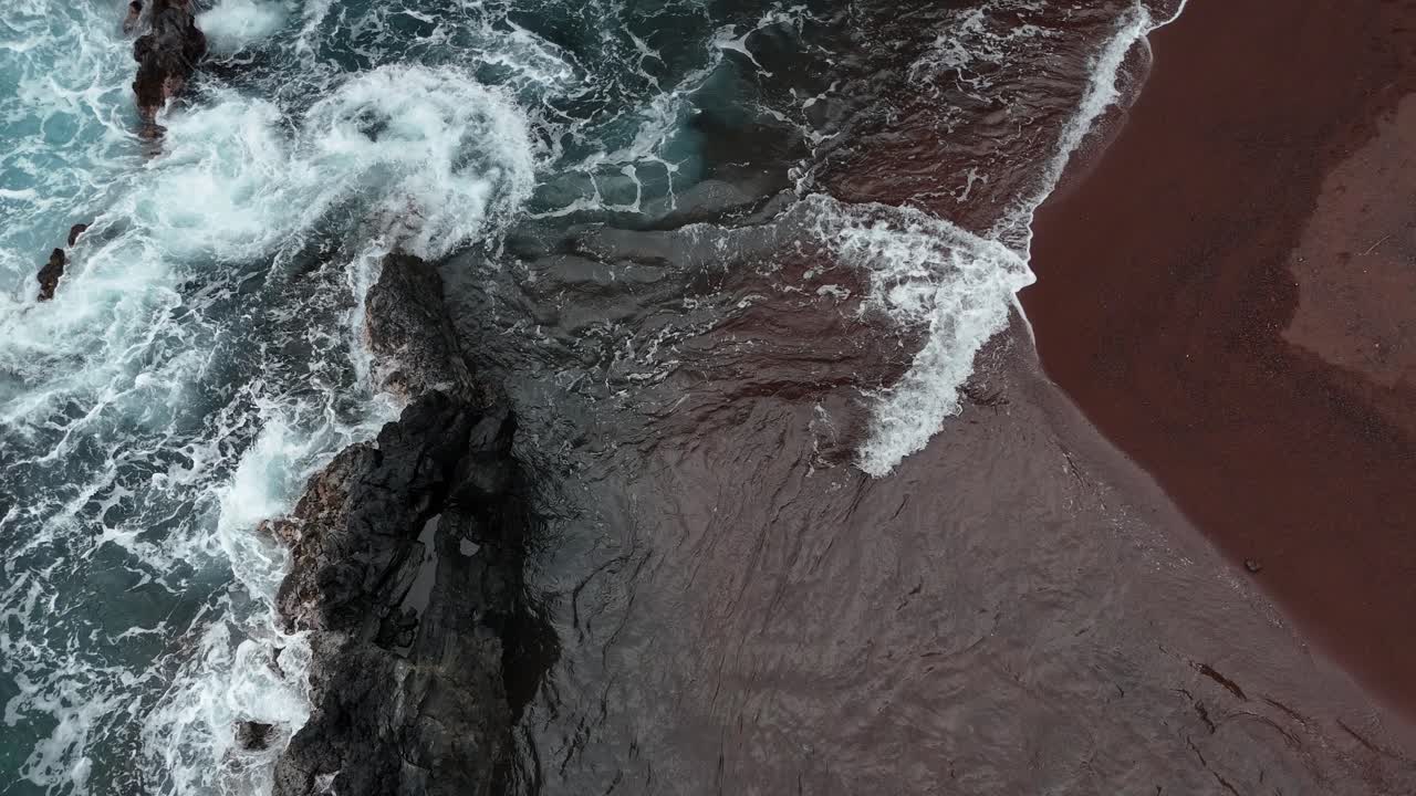 Top-down drone view of waves crashing onto a black rock, creating dramatic white foam patterns in Hawaii