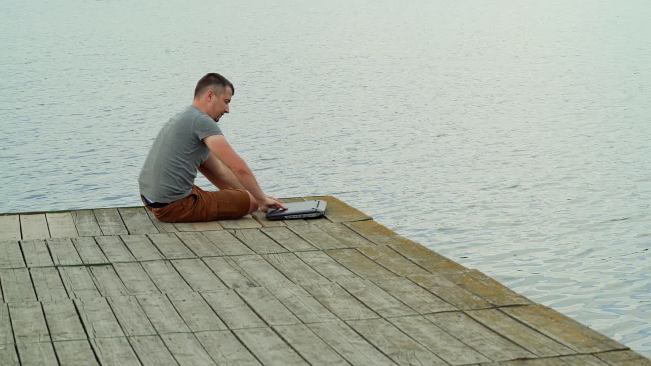 a man is approaching to the end of a masonry and opening a laptop on the background of lake in the summer