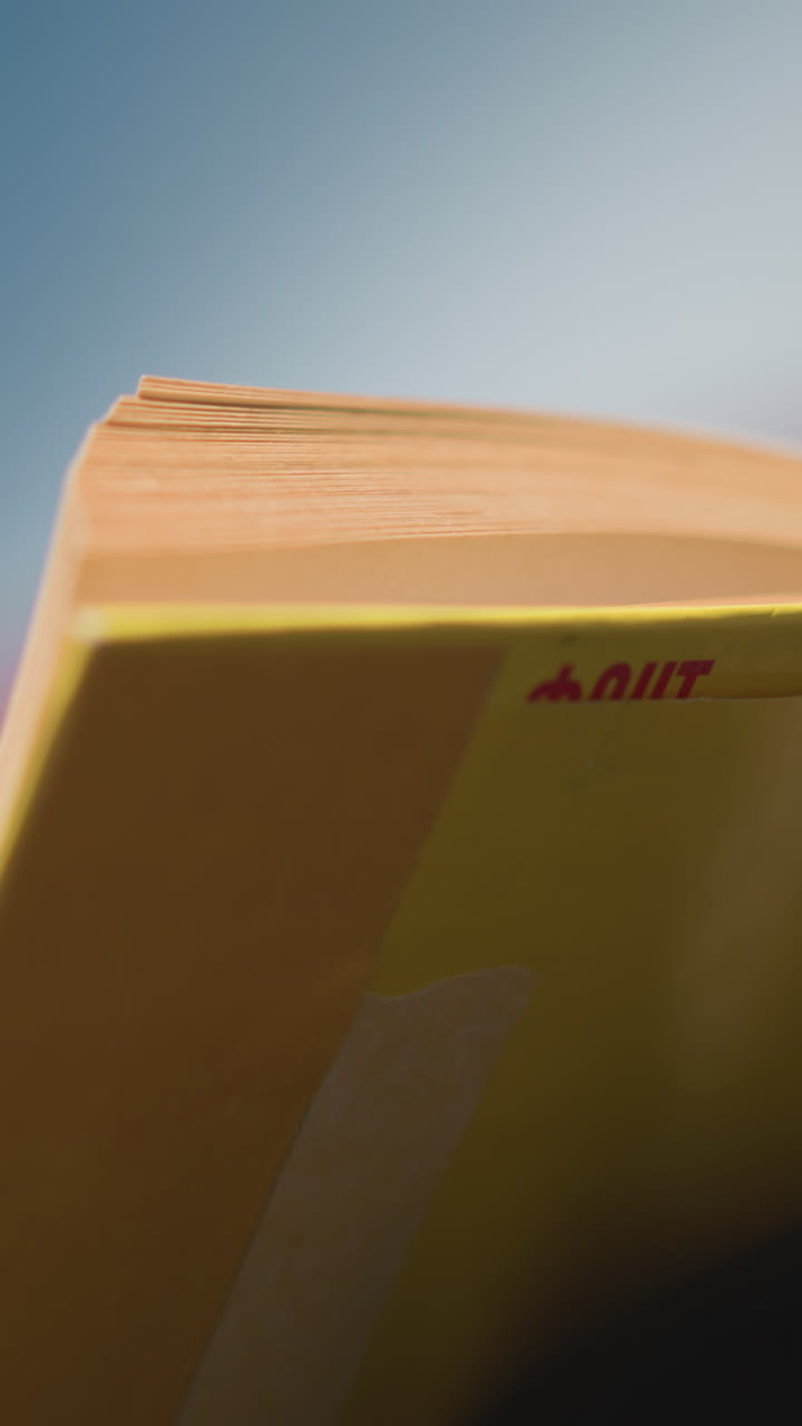 Close-up of lady in outdoor setting, blurred focus on yellow cover book as she flips through the pages, sunlight creating a soft glow, with background softly blurred