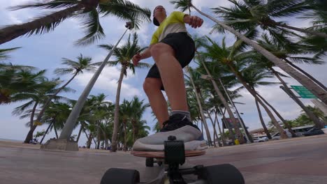 Toma-En-ángulo-Ultrabajo-De-Un-Patinador-Masculino-Cabalgando-Por-La-Playa-Usando-Un-Longboard-Con-La-Vista-De-Palmeras-Tropicales-Detrás-De-él