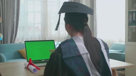 excited asian woman having video call on laptop computer with green mock-up screen. pretty female graduate wearing a graduation gown and cap sitting on the living room