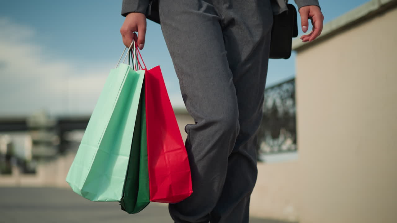 회색 옷을 입은 여성이 오른손에 세 개의 다채로운 쇼핑 가방을 들고 장식 된 울타리 근처에서 고 있으며, 도시 요소와 은 하늘의 흐릿한 배경이 있습니다.