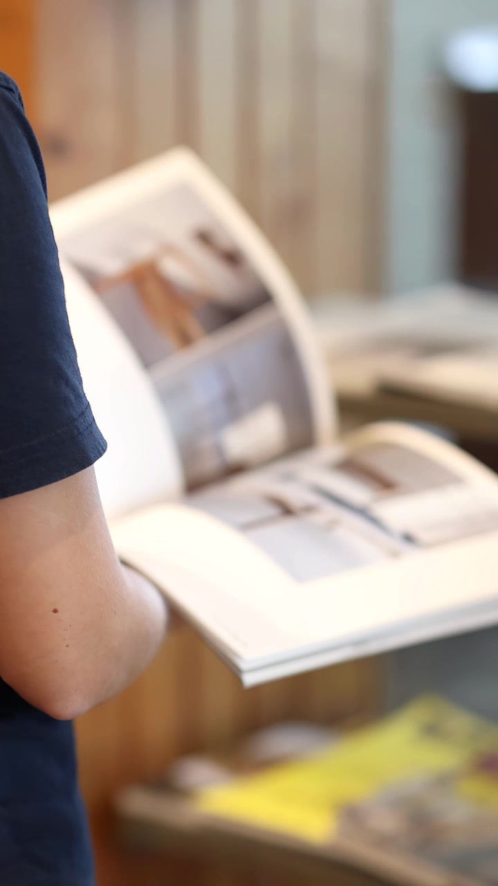 Personne feuilletant et lisant un magazine en intérieur, tenue décontractée, éclairage naturel doux, caméra à main stable.