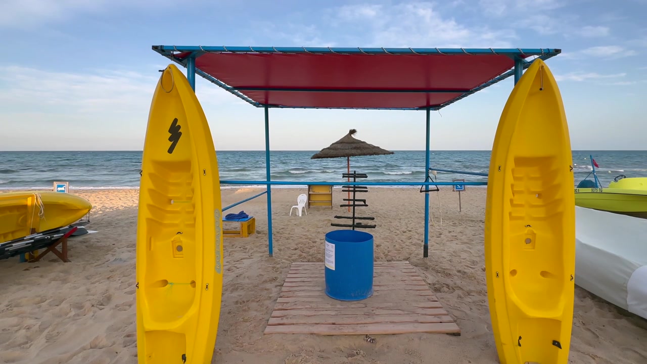 Due kayak giallo brillante sono in piedi accanto a un semplice chiosco di noleggio sulla spiaggia di fronte a un mare calmo, con una luce serale soffusa e un'atmosfera di vacanza tranquilla. Hammamet, Tunisia.