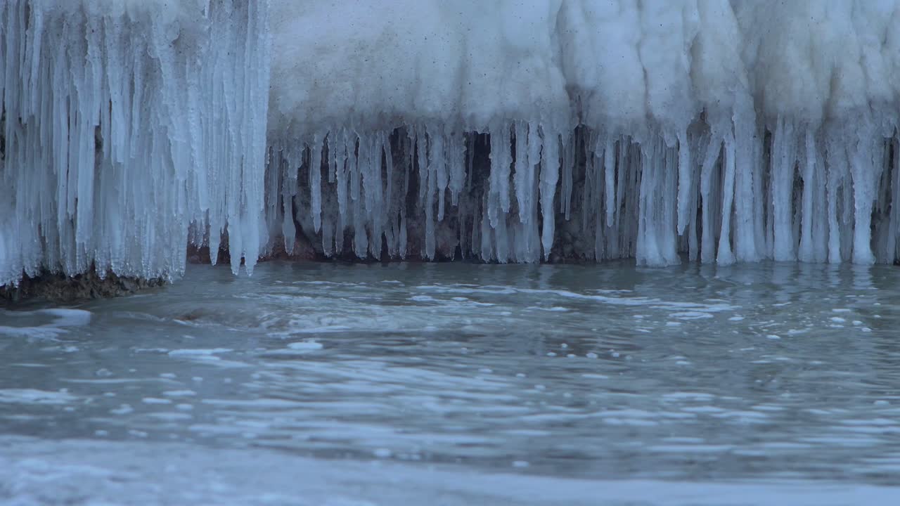 Small waves breaking against the ruins of Karosta Northern Forts fortification on the shore of Baltic sea on a cloudy winter day, covered with ice, snow and icicles, closeup shot