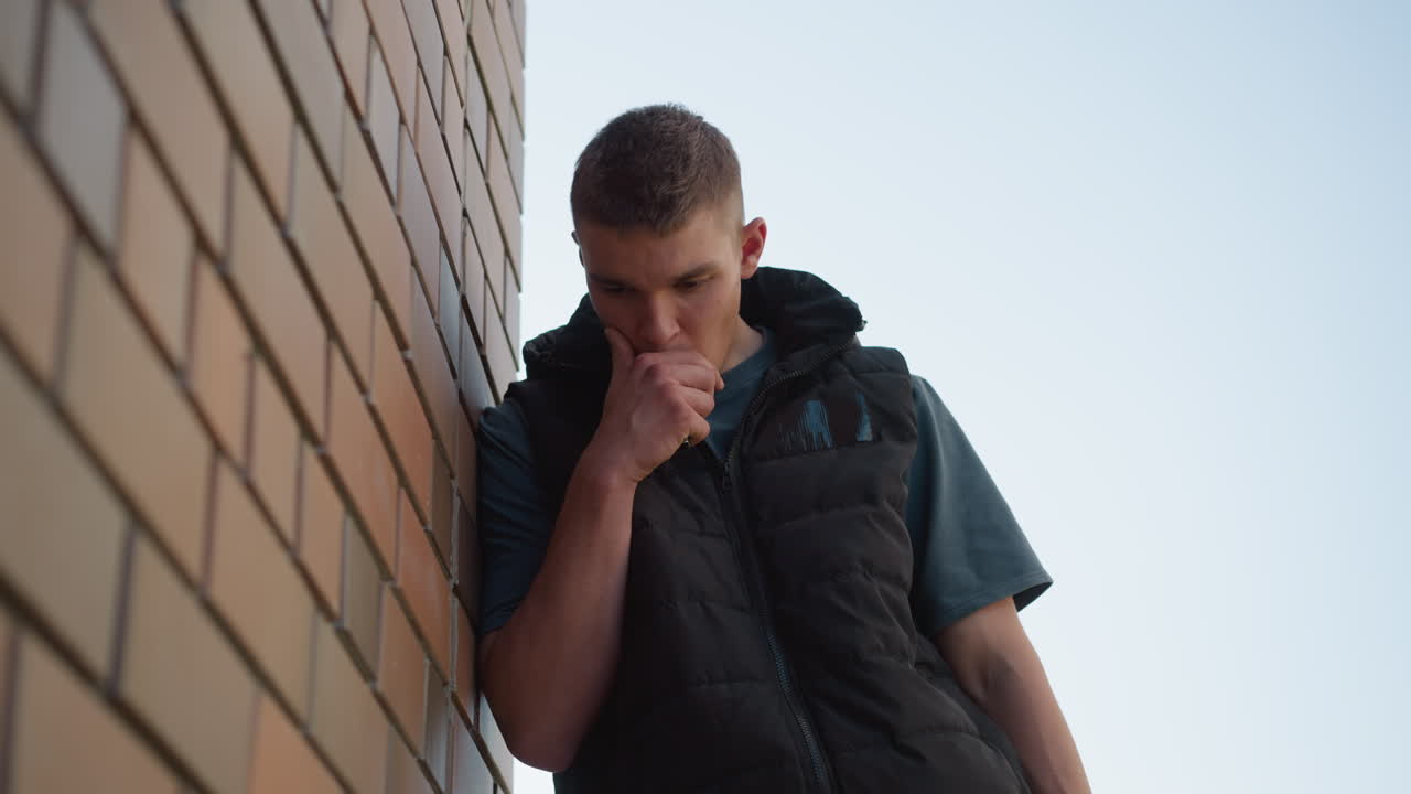 young boy leaning against brick wall outdoors inhaling shisha smoke into mouth as he coughs into hand wearing black puffer vest over dark shirt under soft sunlight casting subtle shadows on wall