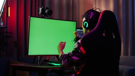 back view of asian boy streamer playing game over network on green screen computer. challenge and bully competitor, live stream video game, desk illuminated by rgb led strip light