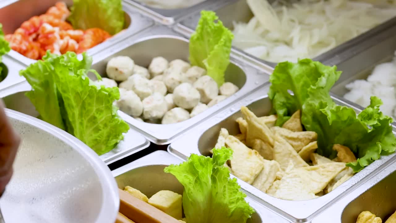 ビュッフェで金属製のトングを使い、豆腐と新鮮な葉物野菜を選んでいる人。