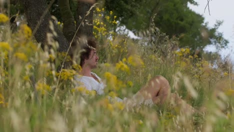 a wide profile shot of a photographer laying under a tree, holding an analog camera, surrounded by tall grass on capri island