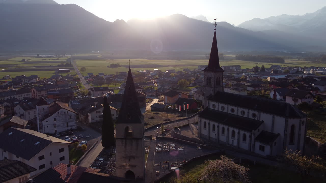 Aerial orbit shot above Vionnaz, Switzerland with two churches illuminated by the sunlight.