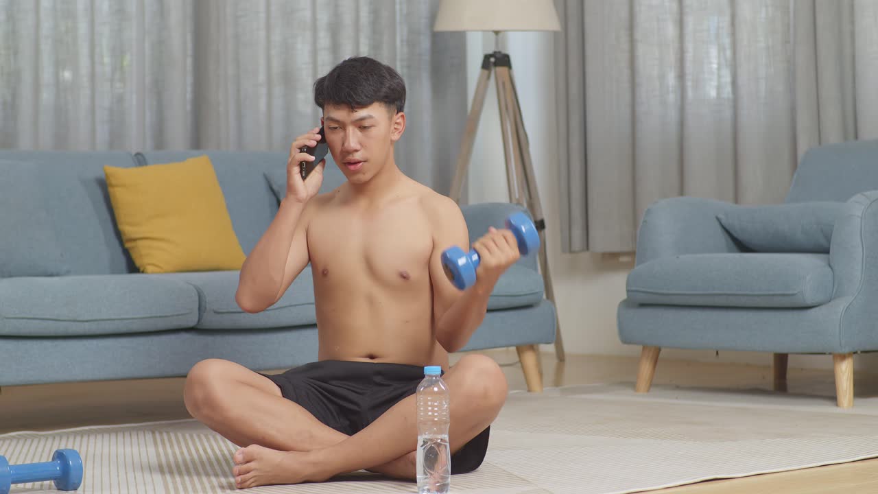 un adolescente asiático entrenando con pesas en la alfombra y hablando en el teléfono inteligente mientras hace ejercicios sin camisa en casa