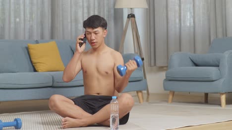 asian teenager boy training with dumbbells on mat and talking on smartphone while doing shirtless workout at home