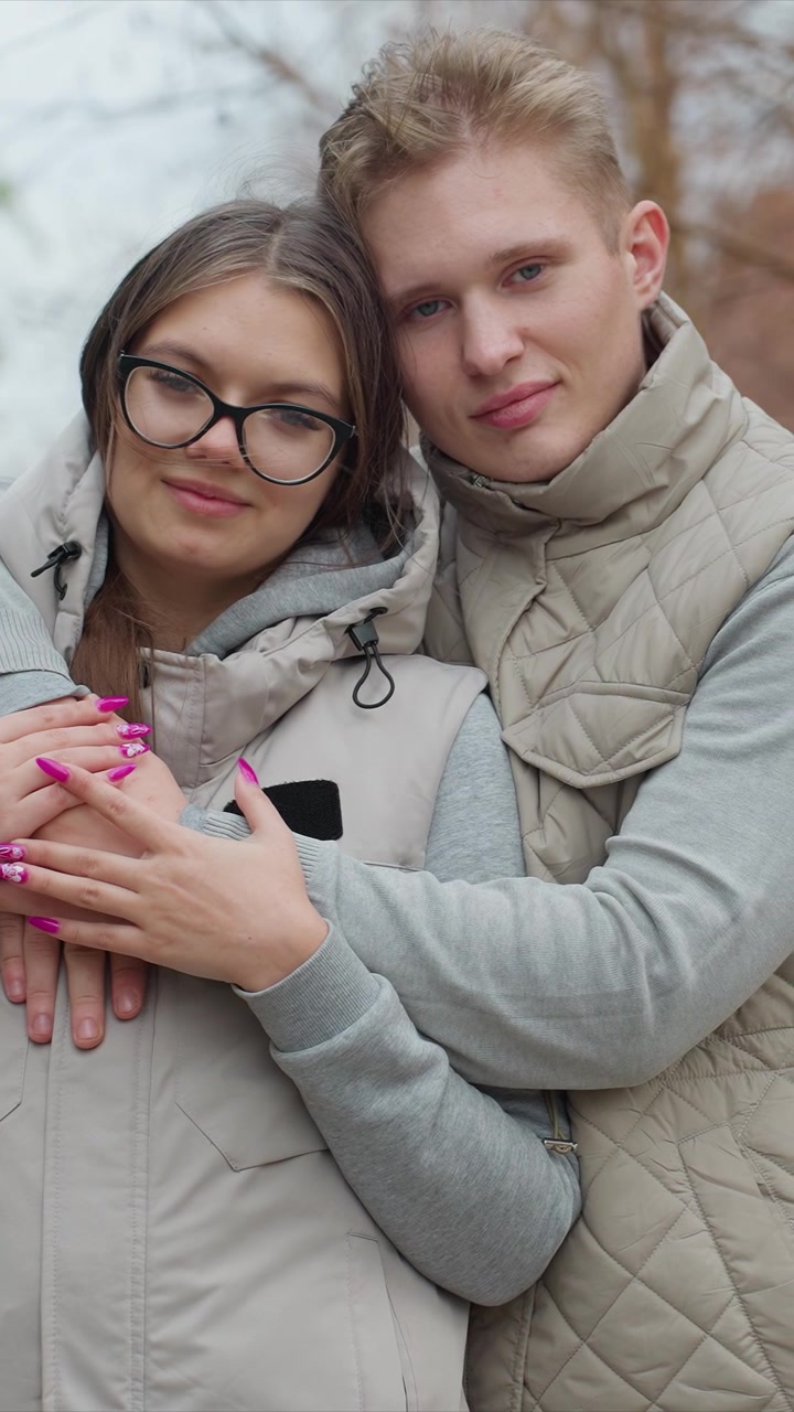 Una donna con smalto rosa abbraccia un'altra persona. Entrambi indossano abiti casual adatti per un ambiente esterno. L'ambientazione include alberi spogli che indicano la stagione autunnale.