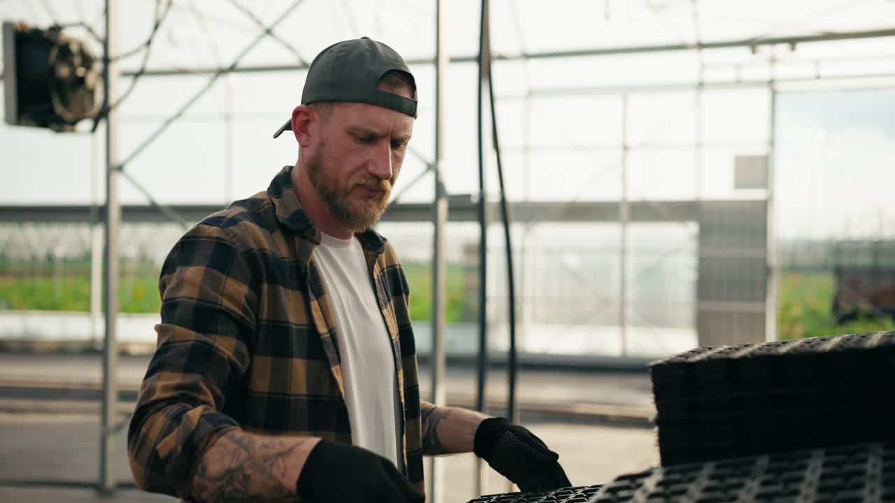 Confident guy Farmer with a beard in a cap in a plaid shirt arranges boxes for seedlings and inspects them during his work in a greenhouse on a farm