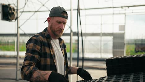 Confident-guy-Farmer-with-a-beard-in-a-cap-in-a-plaid-shirt-arranges-boxes-for-seedlings-and-inspects-them-during-his-work-in-a-greenhouse-on-a-farm