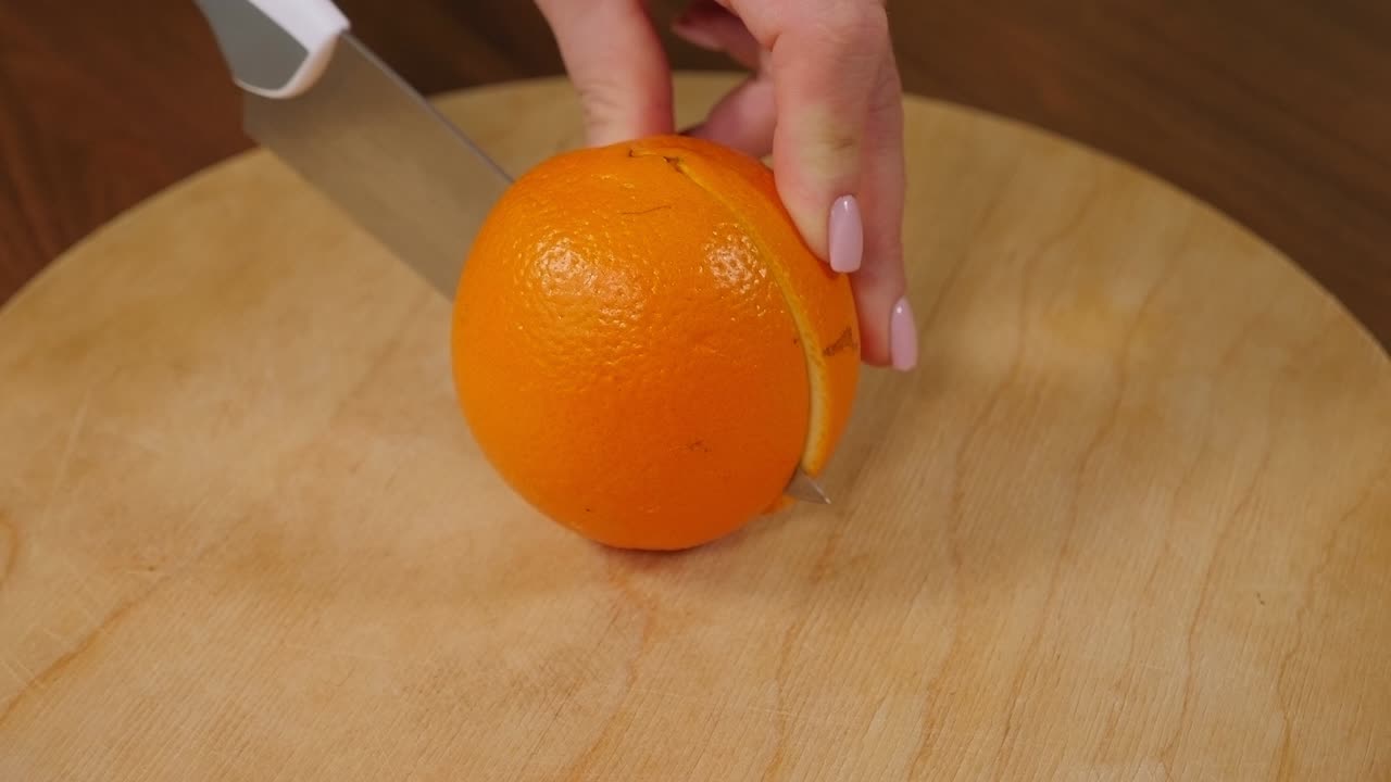 Close-up of a woman cutting an orange in half with a knife on a wooden board.