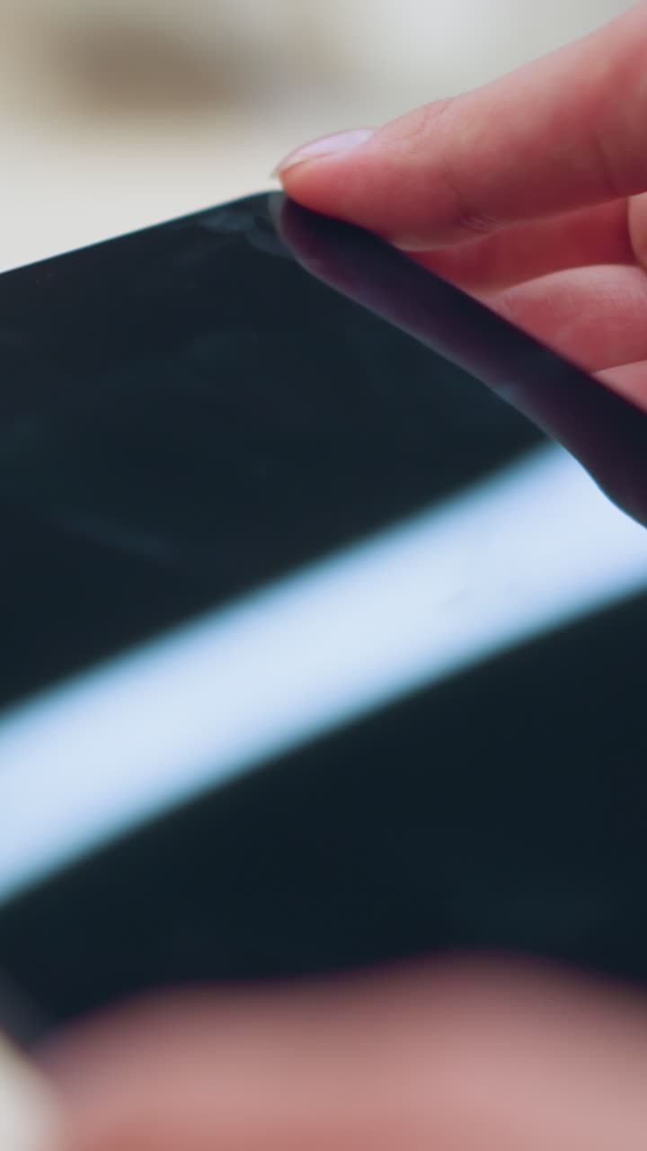 Lady picks up a tablet from its stand in an electronics store, connected by an electric cord, she holds it gently, observing the screen with light reflecting off it with the background well lit up