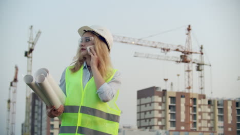 Portrait-of-a-woman-engineer-in-a-helmet-talking-on-the-phone-on-the-background-of-construction-with-cranes-holding-drawings-in-his-hands.-Female-engineer-on-construction-site.