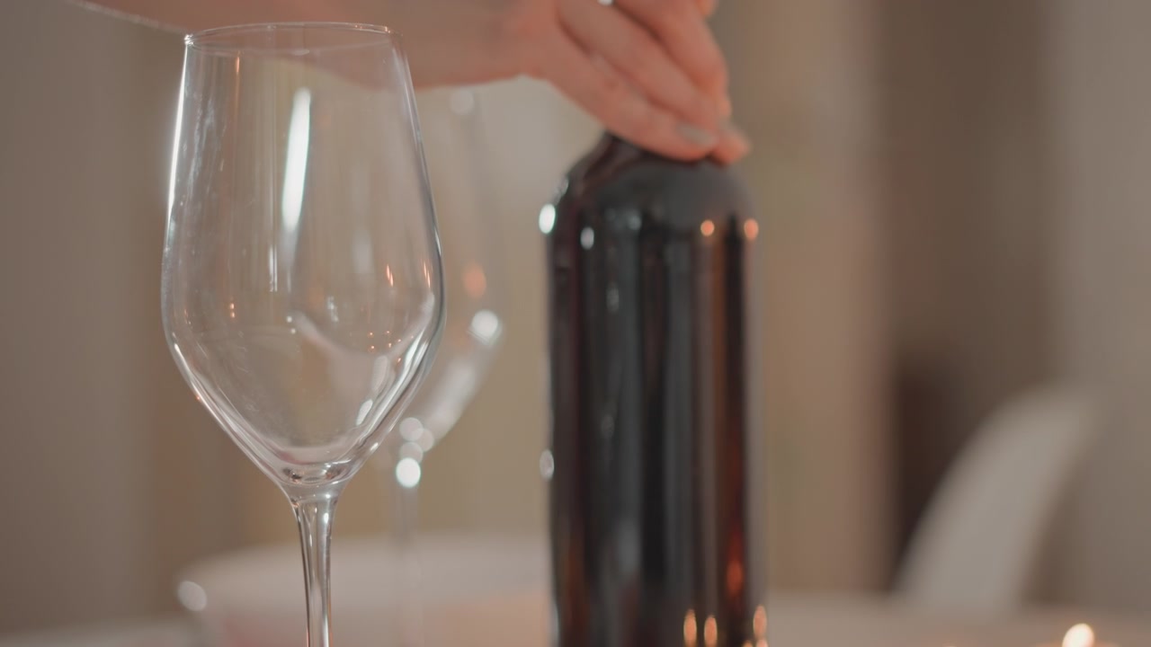 Nærbilde av et tomt vinglass i forgrunnen med en mørk flaske bak på bordplaten, mykt innendørs lys, svak uskarphet og dråper inne i glasset antyder nylig bevegelse, en varm og elegant spiseatmosfære.