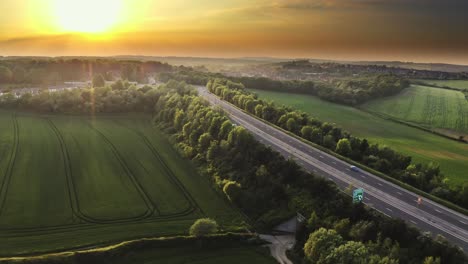 Vista-Aérea-Sobre-Una-Carretera-Rodeada-De-Campos-Verdes-Durante-La-Puesta-De-Sol-En-El-Reino-Unido