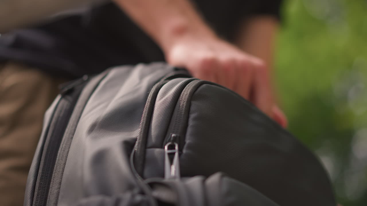 White Man Unzipping Backpack Outdoors In Park, Hands Closeup On Gray Nylon Bag Zipper, Focused Retrieval Of Laptop And Travel Gear, Warm Daylight And Green Blurred Background, Casual Clothing