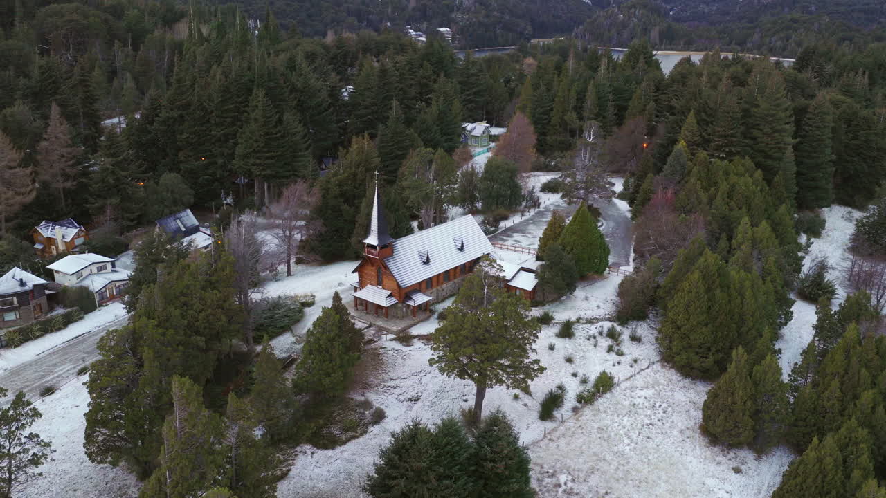 Widok z lotu ptaka na małą drewnianą kaplicę w zaśnieżonej leśnej osadzie w Bariloche, ukazujący spokojny zimowy styl życia