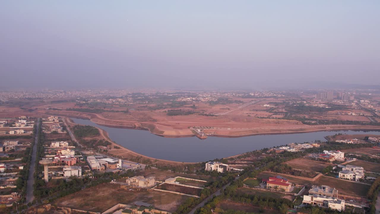 Rekaman udara luas komplek perumahan Gulberg Green yang menampilkan danau besar dan arsitektur modern saat matahari terbenam yang berkabut