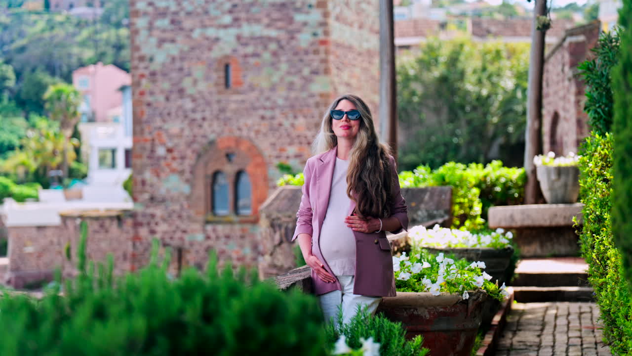 Pregnant woman with long hair, wearing sunglasses standing with the Chateau de la Napoule Castle in Mandelieu-La Napoule, France in the background