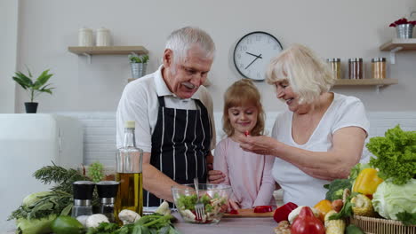 Senior-couple-grandmother-and-grandfather-in-kitchen-feeding-granddaughter-child-with-chopped-pepper