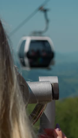 female tourist adjusts modern tower viewer on point at cable way station against foggy old mountains close backside view slow motion