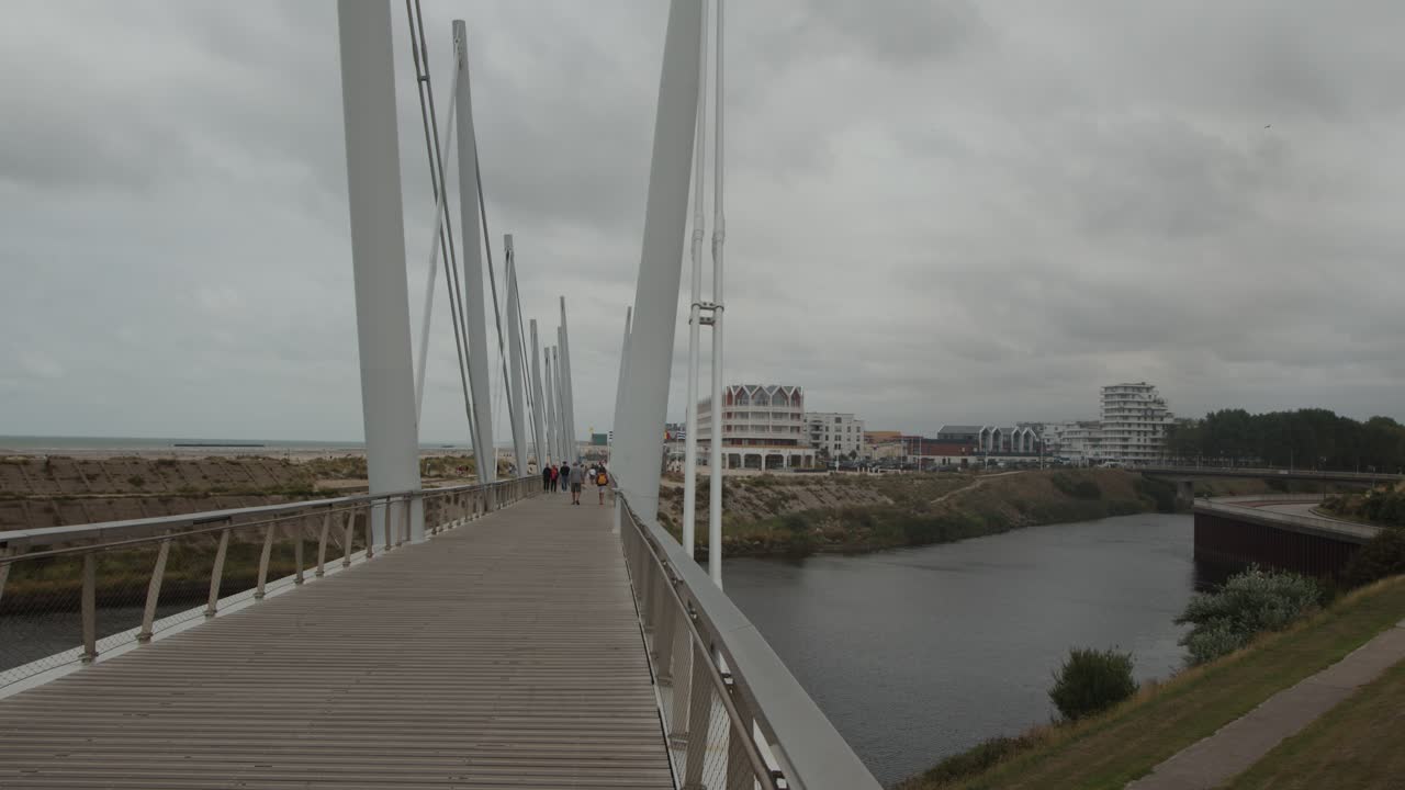 Steady camera moves along contemporary bridge over river, cloudy sky, urban waterfront, daytime lighting
