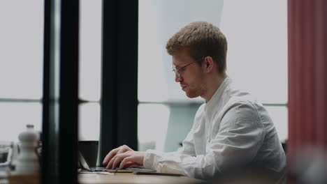 Un-Especialista-Independiente-En-TI-Está-Trabajando-Con-Una-Computadora-Portátil-En-Una-Cafetería-Durante-El-Día.-Retrato-De-Un-Hombre-Con-Un-Cuaderno.