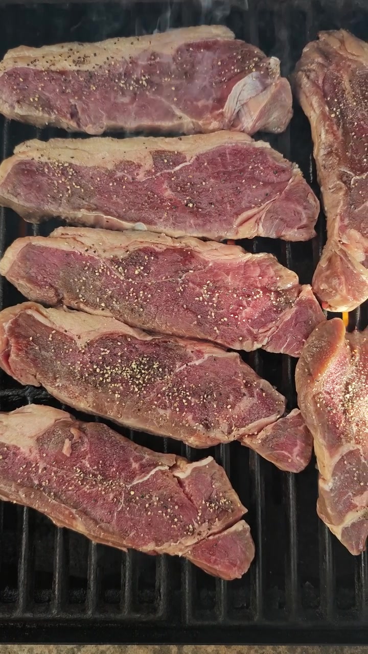 Scatto verticale dall'alto di bistecche di controfiletto all'inizio della grigliatura, leggermente condite con sale e pepe su una griglia calda, carne cruda che tocca appena la griglia, prima scottatura, processo culinario pulito