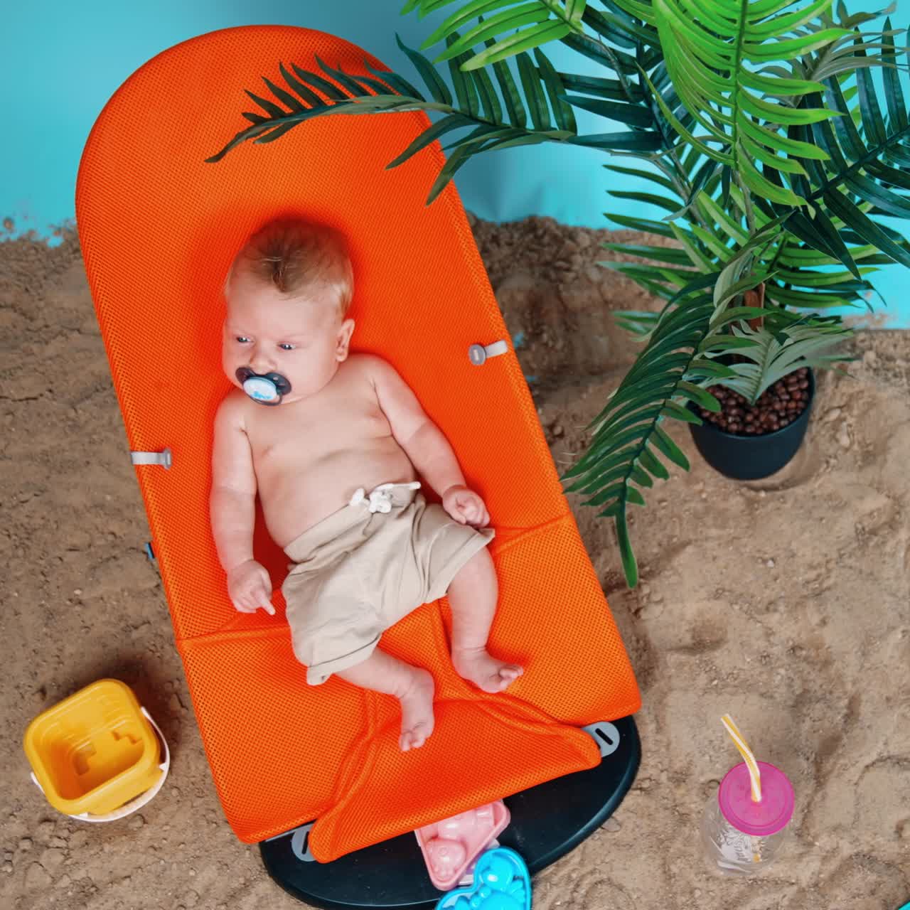 Sweet Caucasian baby boy wearing shorts lies peacefully in the chair. Beach sand and pot with plant nearby. Top view