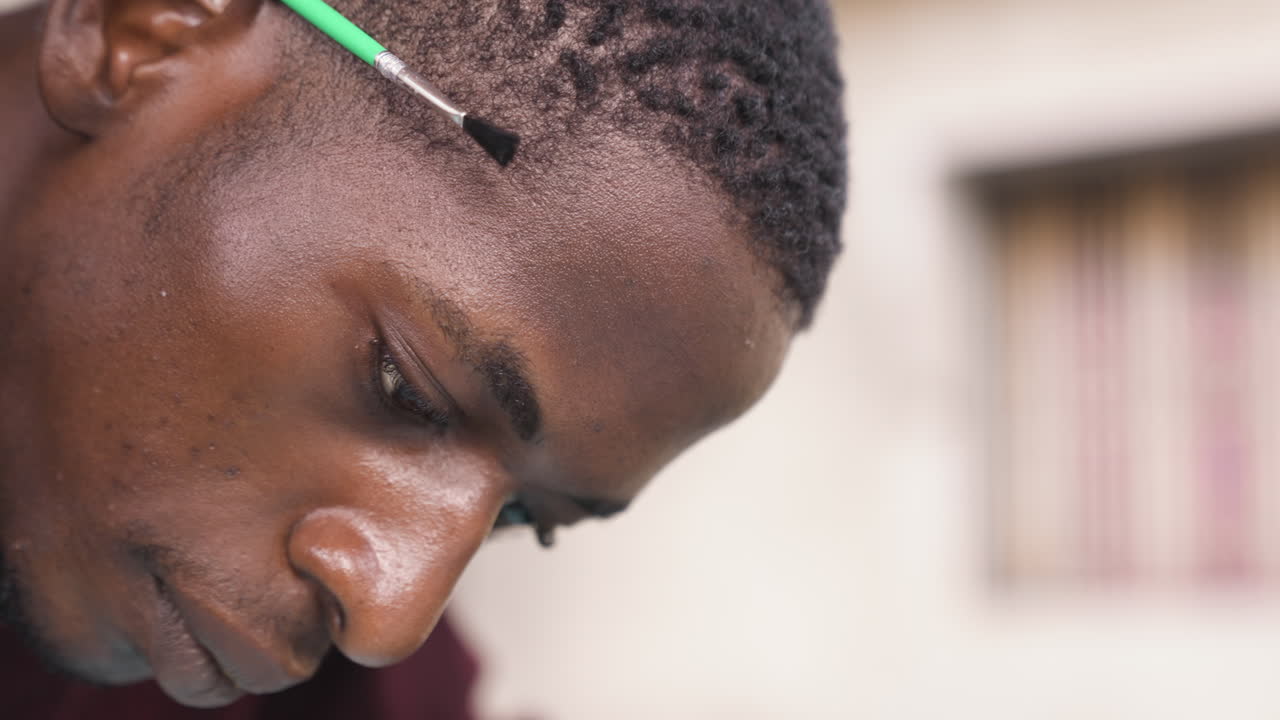 Primo piano di un uomo di colore con un pennello che applica colore sull'attaccatura dei capelli, punta del pennello e pigmento visibili, cuoio capelluto texturizzato e sudato, profondità di campo ridotta, luce da studio che evidenzia i tratti e la tecnica deliberata