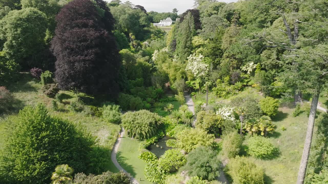 White mansion sitting at head of green tree lined tropical garden paradise with pathways on valley floor. Summer day.