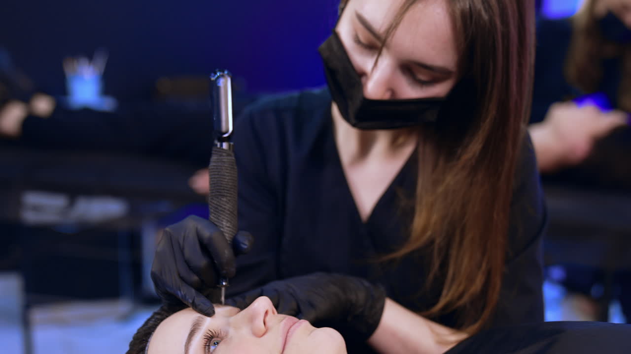 Brow-maker in black mask and gloves uses special tool for permanent make up. Client is having salon procedure on eyebrows. Close up.