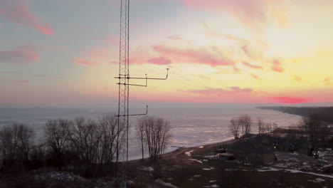 Ein-Windmesser,-Der-Auf-Einem-Turm-Weht,-Mit-Einem-Wunderschönen-Orangefarbenen-Und-Rosafarbenen-Sonnenuntergang-Und-Einem-Gefrorenen-Wintersee-Im-Hintergrund