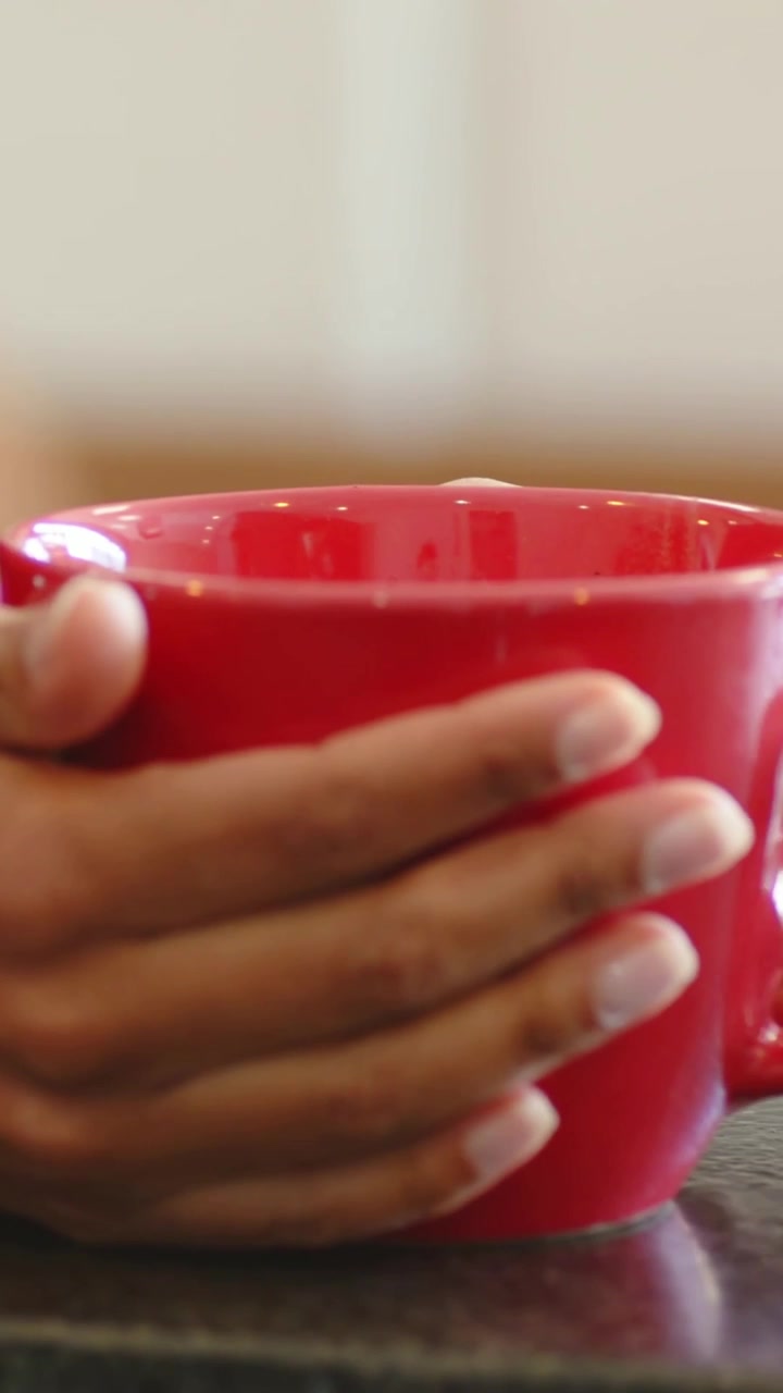 Vertical video: Mug appearing, female hand grabbing red mug at kitchen table in magenta top to sip