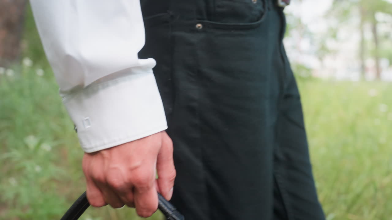 Close leg view of man in black trousers and white shirt carrying black leather bag while walking slowly along green forest path surrounded by grass and trees during summer outdoor exploration
