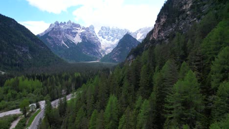 Sideway-aerial-shot-over-forest-covered-mountain-valley-reveals-snowy-Mount-Cristallo,-Dolomiti,-Italy