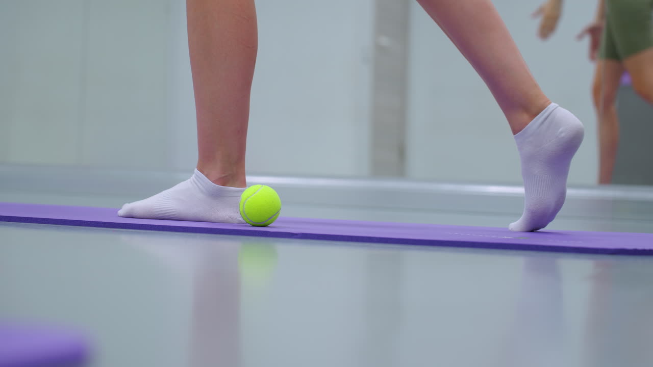 vista lateral de la pierna de un ejercicio de fitness en una alfombra púrpura con una pelota de tenis verde cerca, el espejo refleja a la persona estirándose hacia adelante, mostrando el equilibrio y el movimiento controlado en un entorno de gimnasio activo