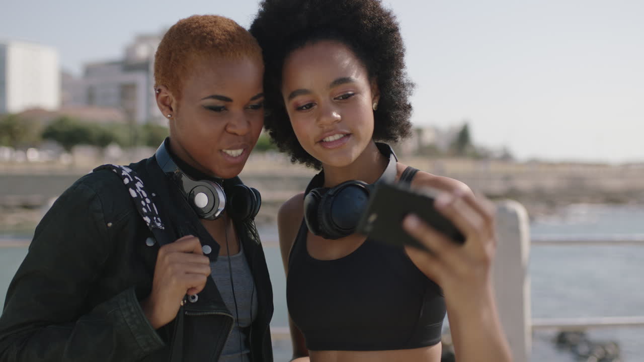 portrait of two beautiful african american woman uing phone on beachfront posing taking selfie